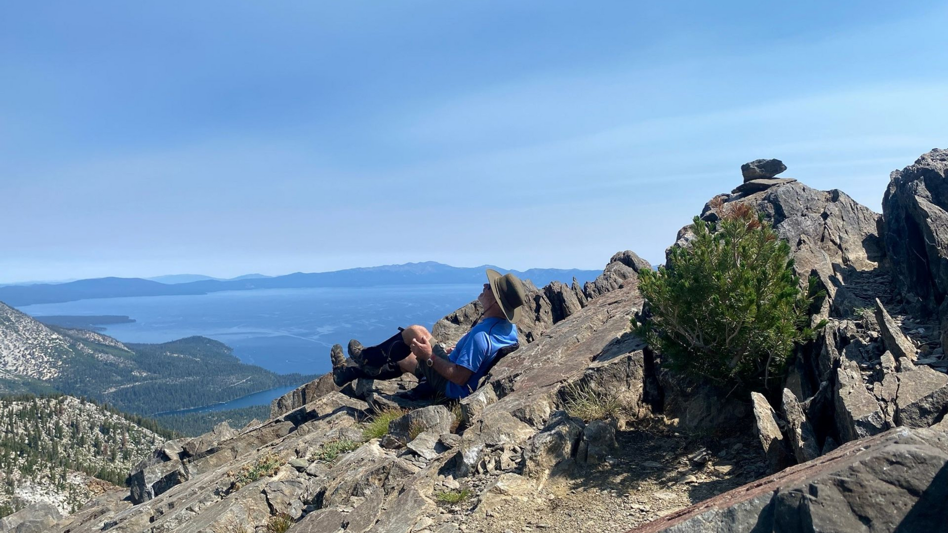 Person resting on a rocky mountain peak, overlooking a blue sea and islands on a sunny day.