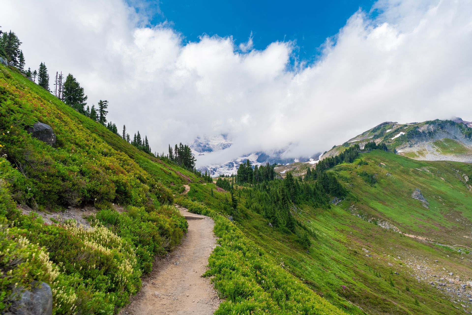 A hiking trail winds along a lush, green mountainside, with a cloudy sky overhead.