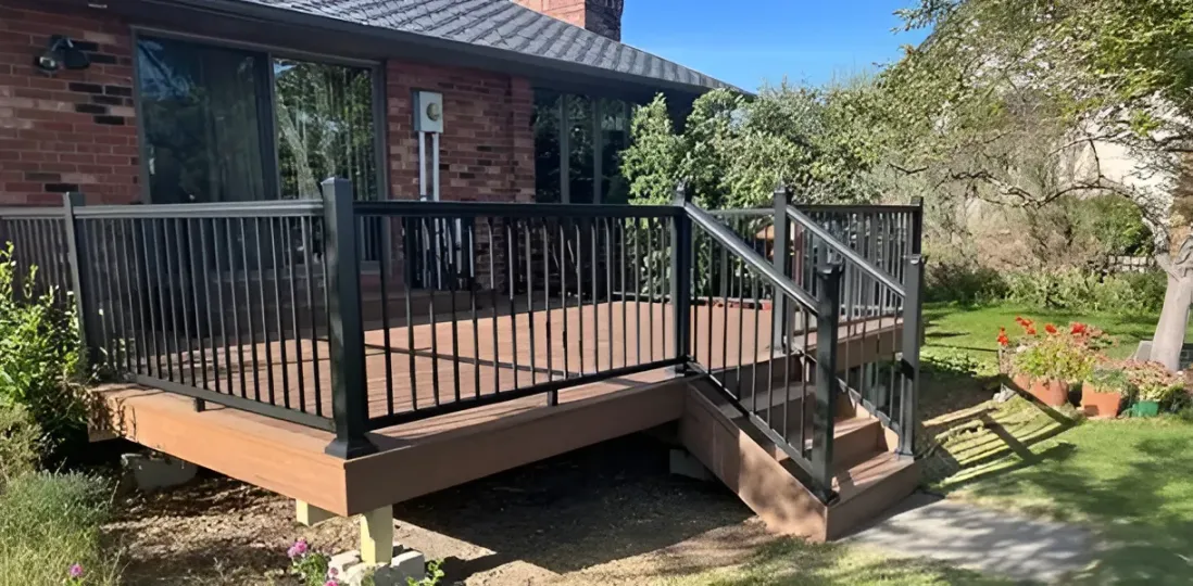Wooden front deck with black railing and stairs beside a brick house, overlooking a sunny garden