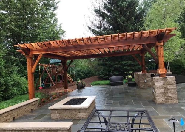 Wooden pergola over a stone patio with a fire pit, surrounded by trees and outdoor seating