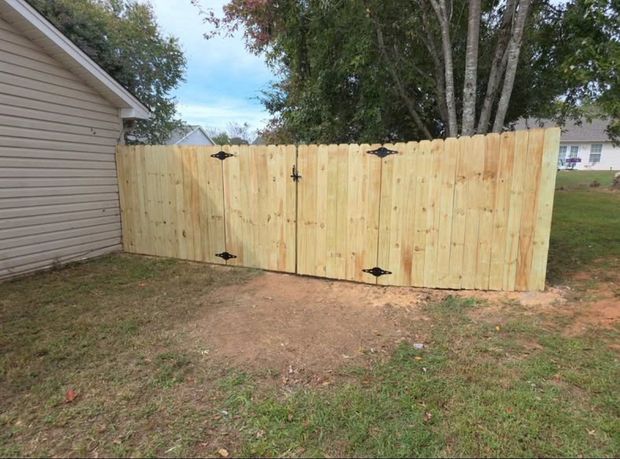 New wooden fence gate in a yard beside a house, with dirt and grass in front