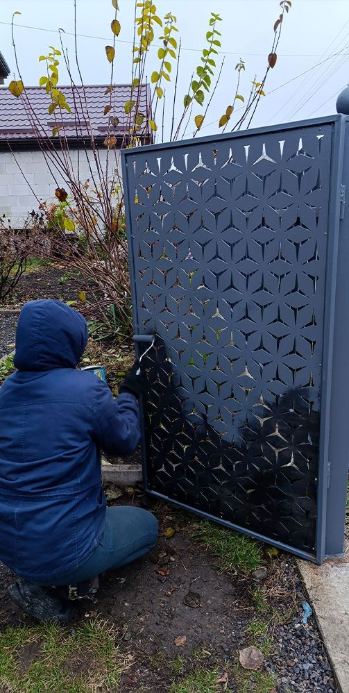 Person in blue jacket painting a dark decorative metal fence outdoors in a garden.