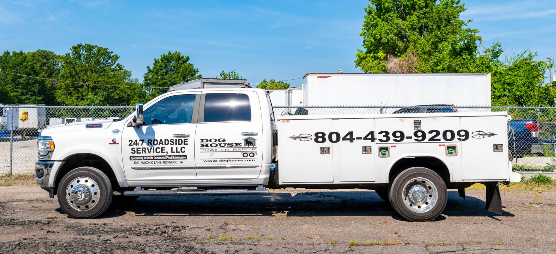White utility truck with service markings parked on a gravel lot under a blue sky, with trees behind it