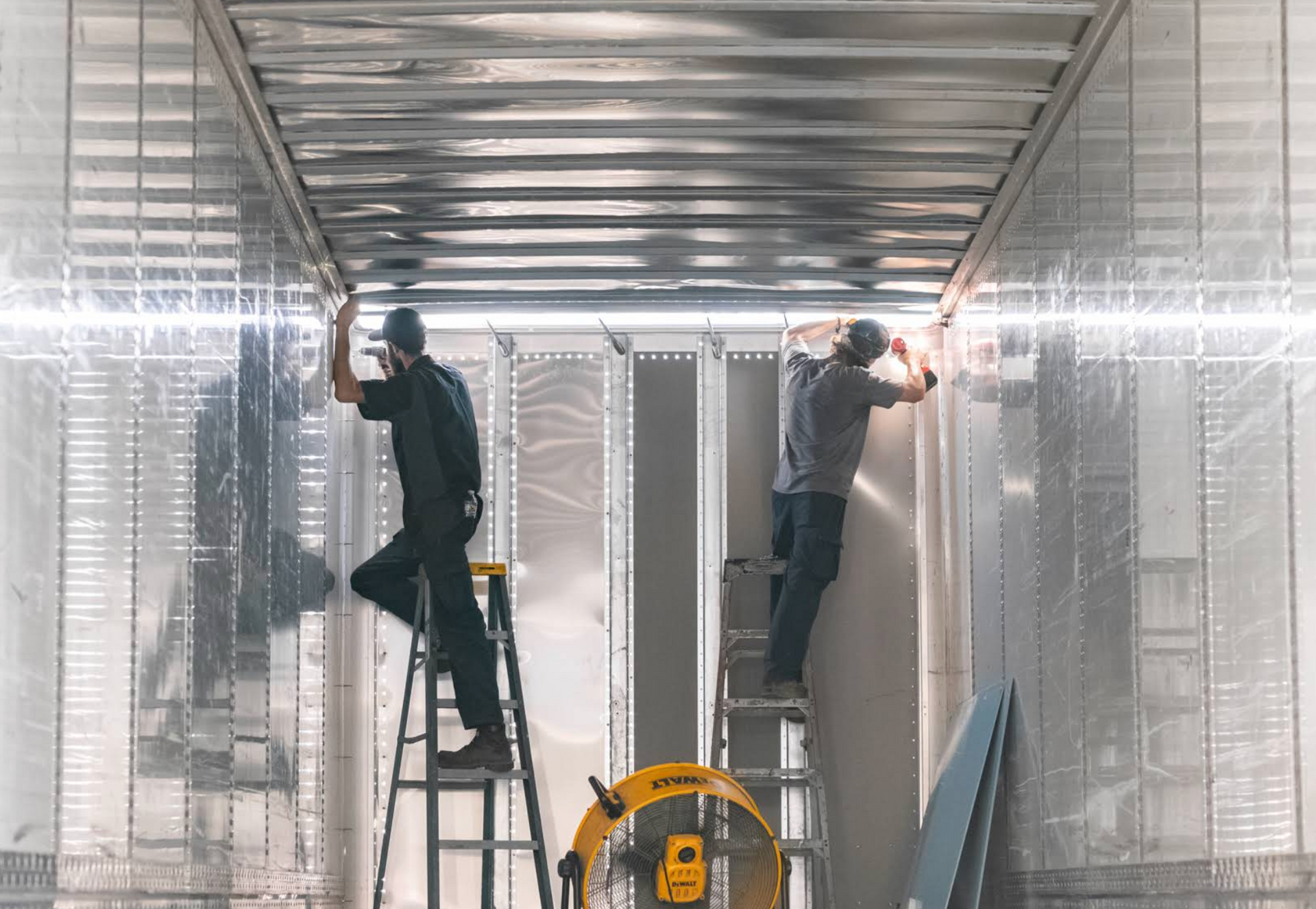Two workers on ladders installing a metal ceiling in a bright industrial room
