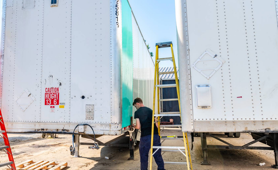 Worker inspecting equipment between two white trailers, with a yellow ladder in the center outdoors.