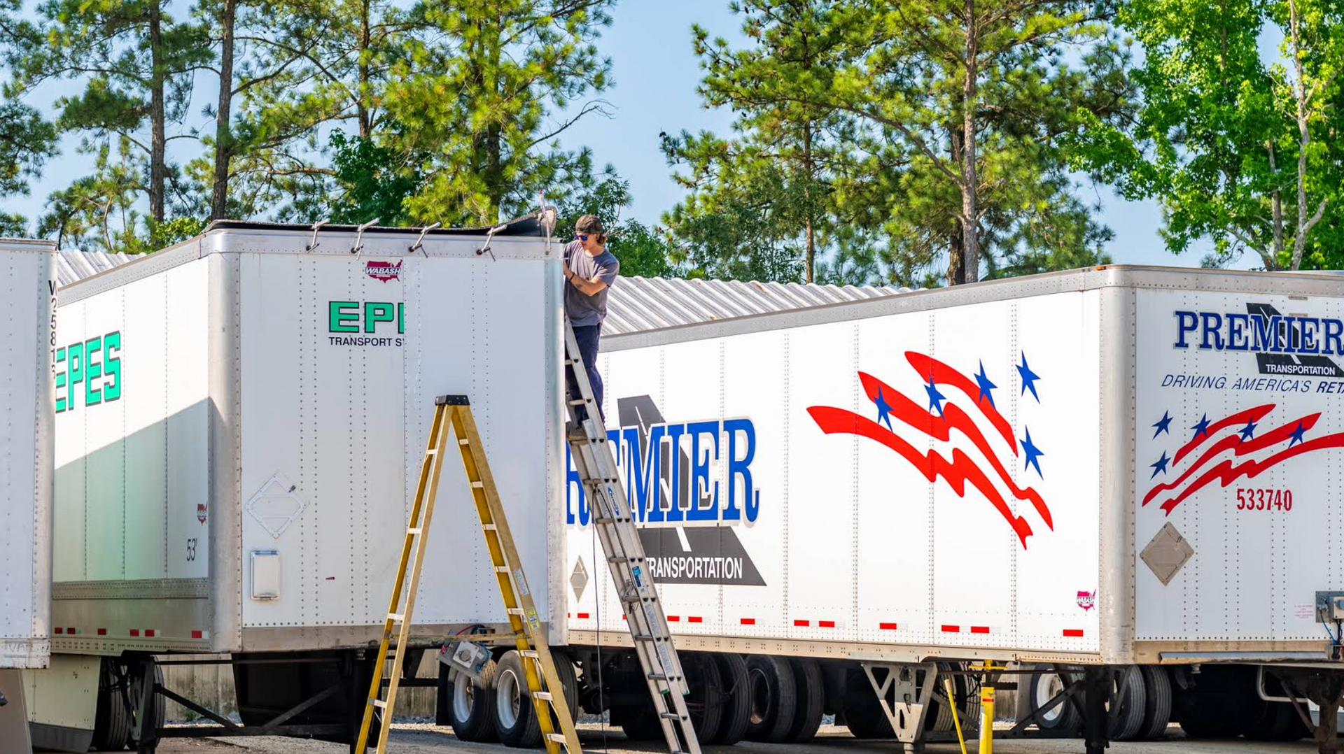 Two white semi-trailers parked outdoors, one with ladders and colorful logos, against trees and blue sky
