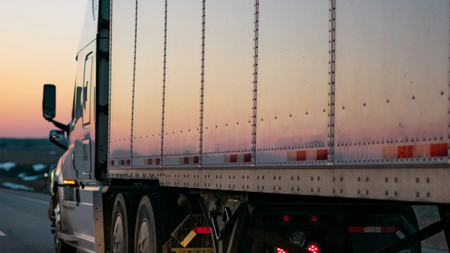 Semi-truck trailer on a highway at sunset, with pink-orange sky reflected on its side