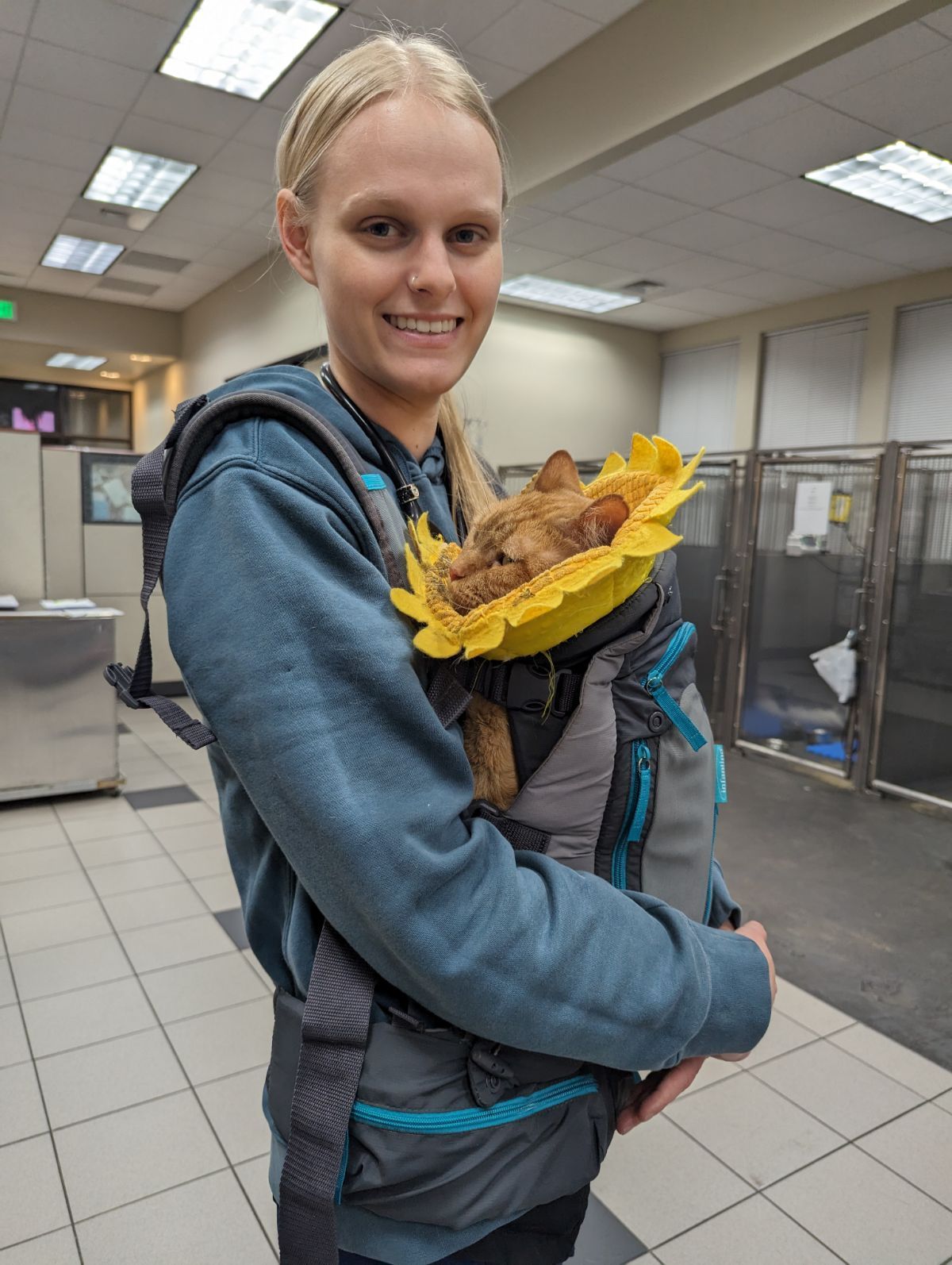 Person holding a small dog wrapped in a yellow blanket, smiling in an indoor shelter hallway.