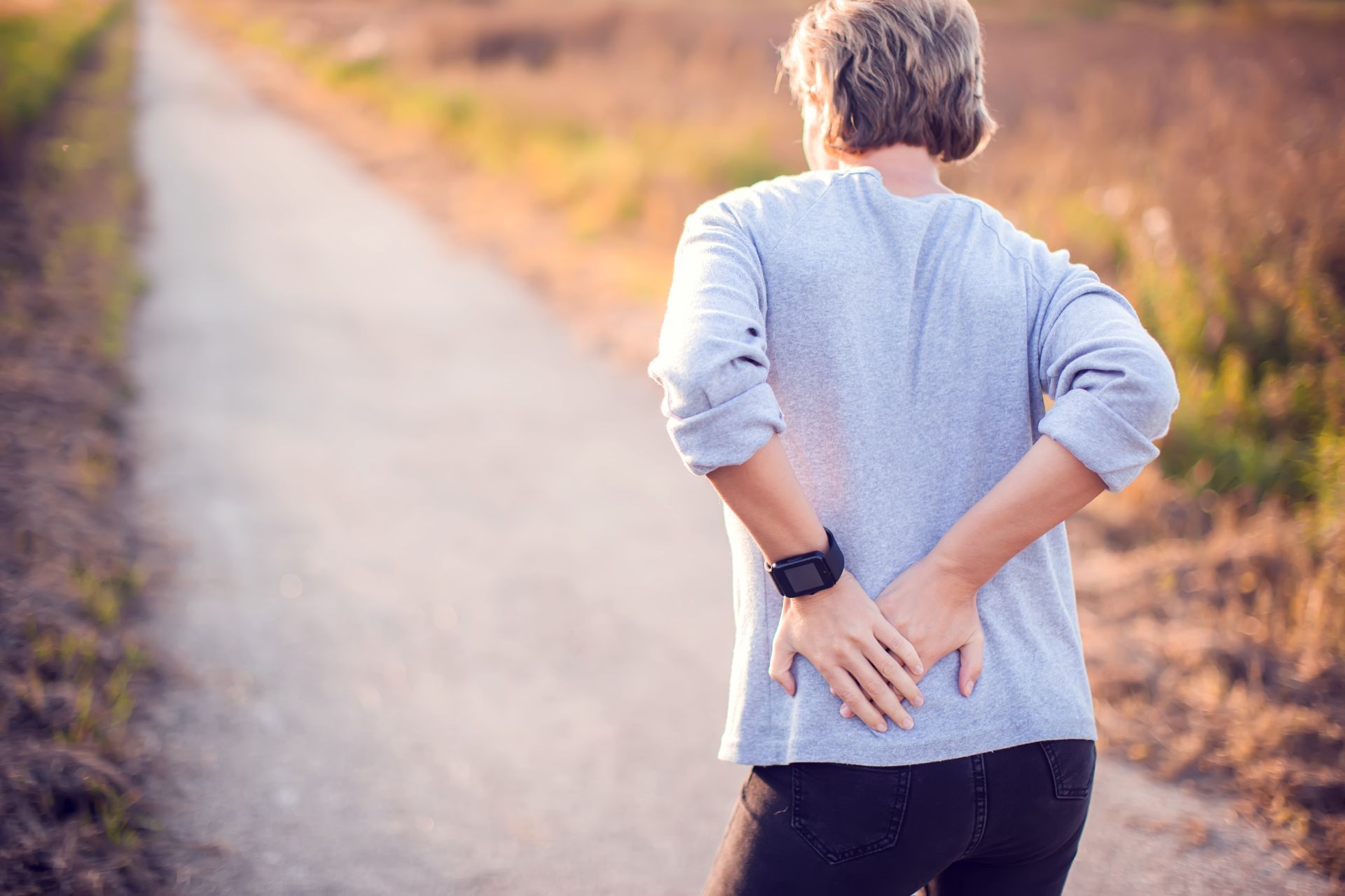 Person in a light blue shirt standing on a path, reaching behind to hold their lower back.