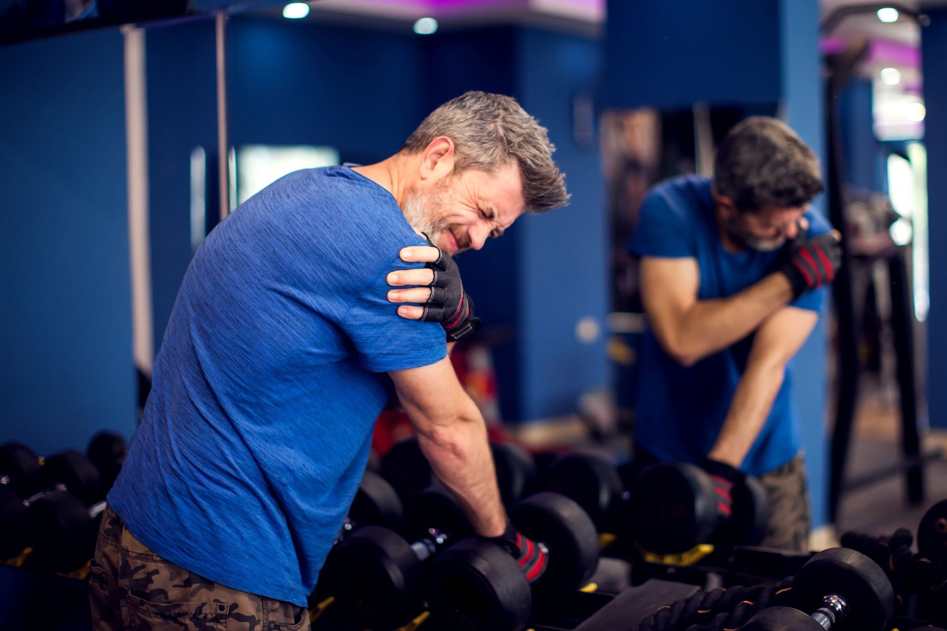 Man in blue shirt leaning over dumbbells in a gym, checking his shoulder in a mirror.