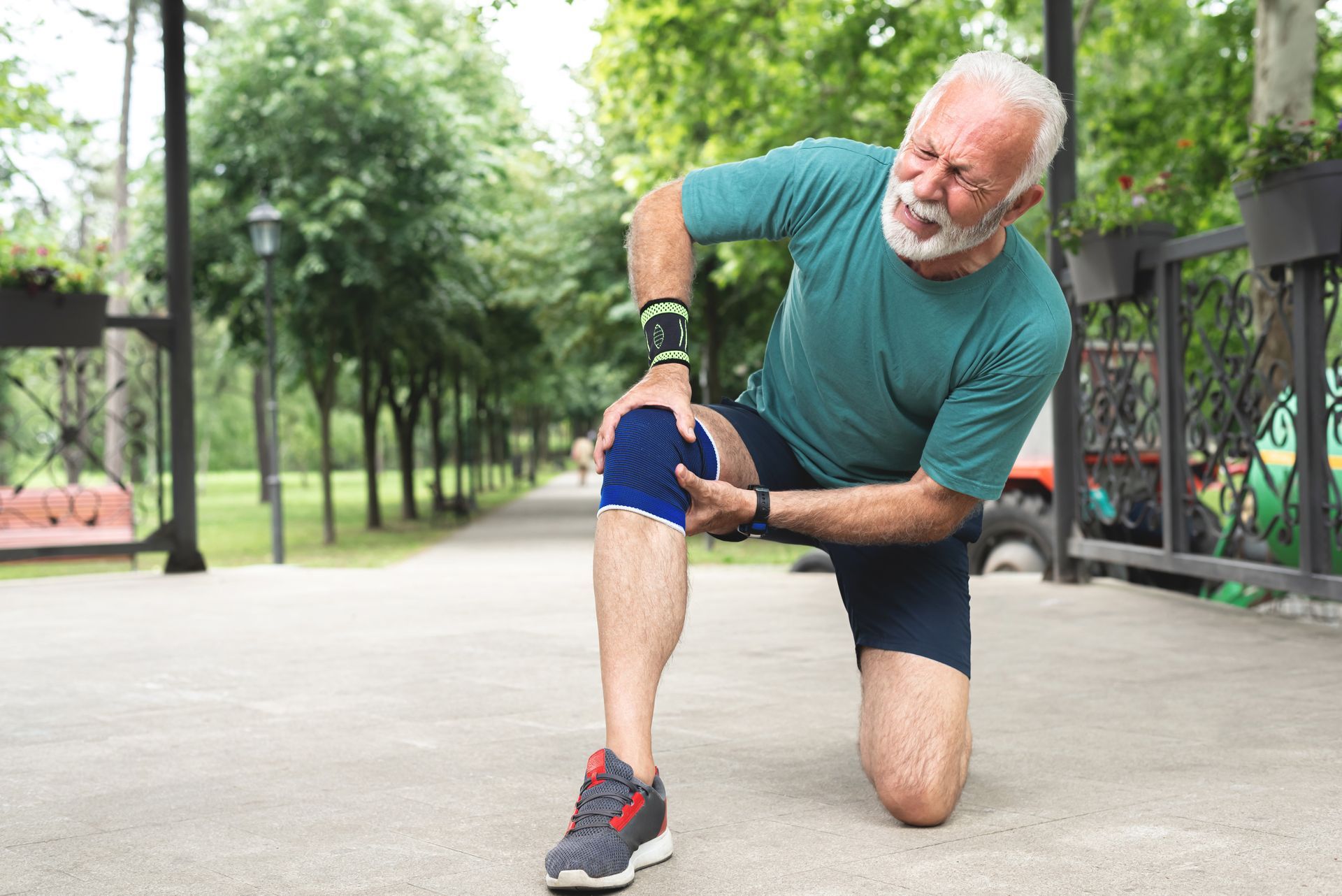 Man kneels on a park path, holding his knee in pain while wearing a blue sleeve.