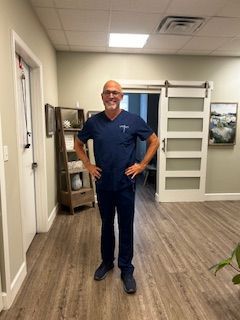 Man in navy scrubs standing in a hallway with hands on hips, near a doorway and wall art.