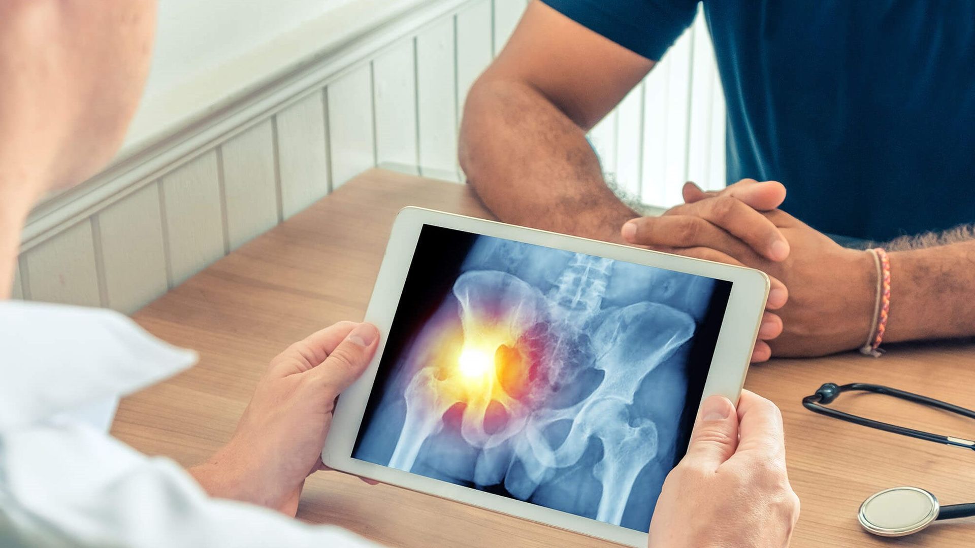 Hands holding a tablet showing a glowing hip X-ray, with a doctor at a desk nearby.