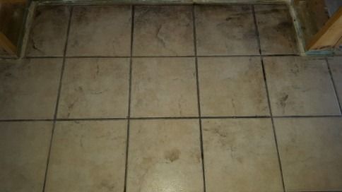 Dirty beige tiled floor with dark grout lines and scattered stains near a doorway