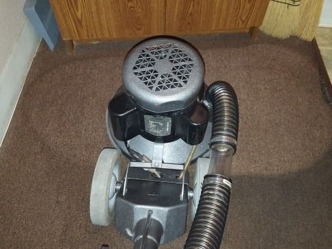 Grey vacuum cleaner on a brown carpet near a wooden cabinet and curtain.
