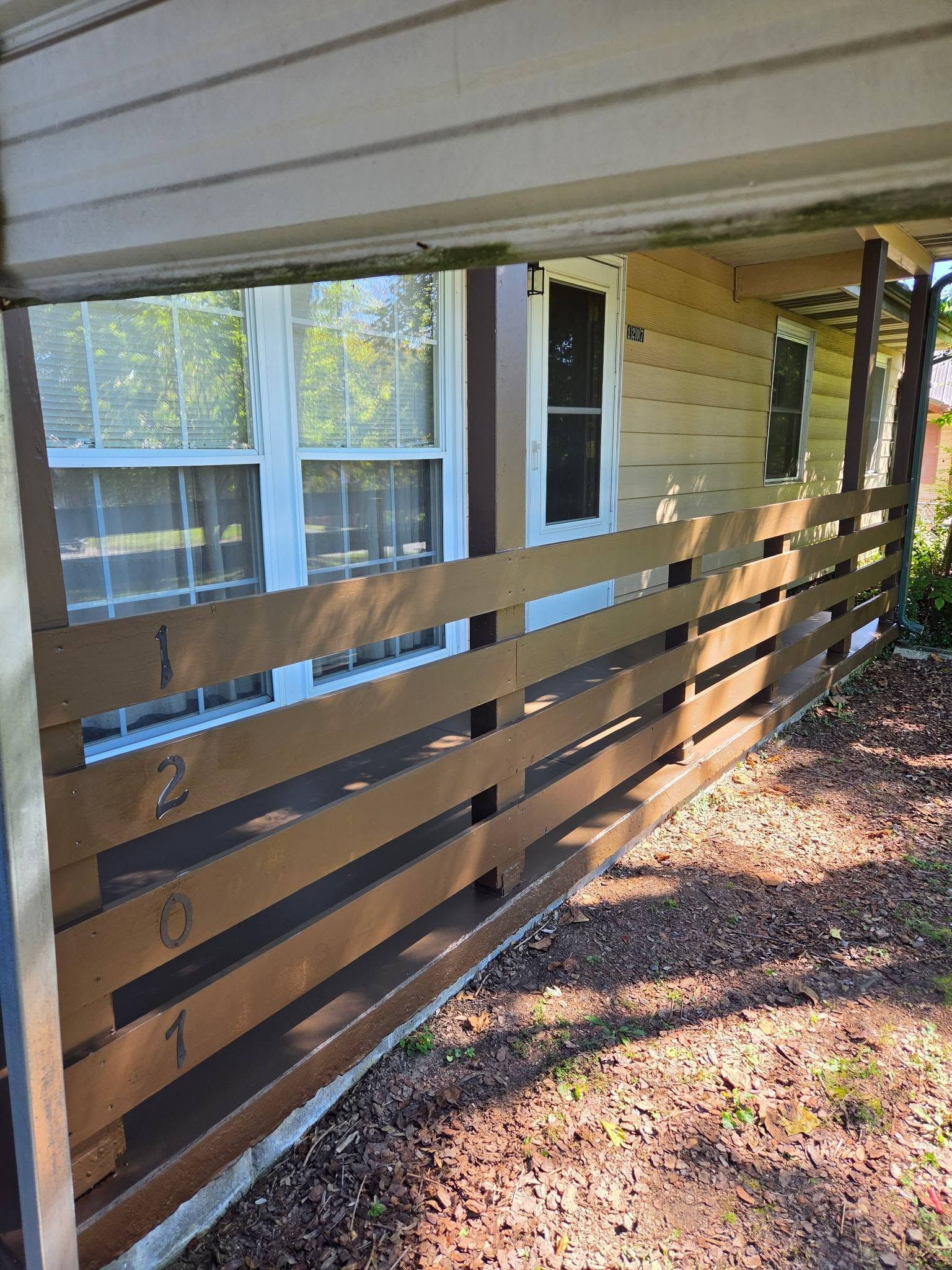Brown slatted porch railing beside beige house with windows and a shaded walkway