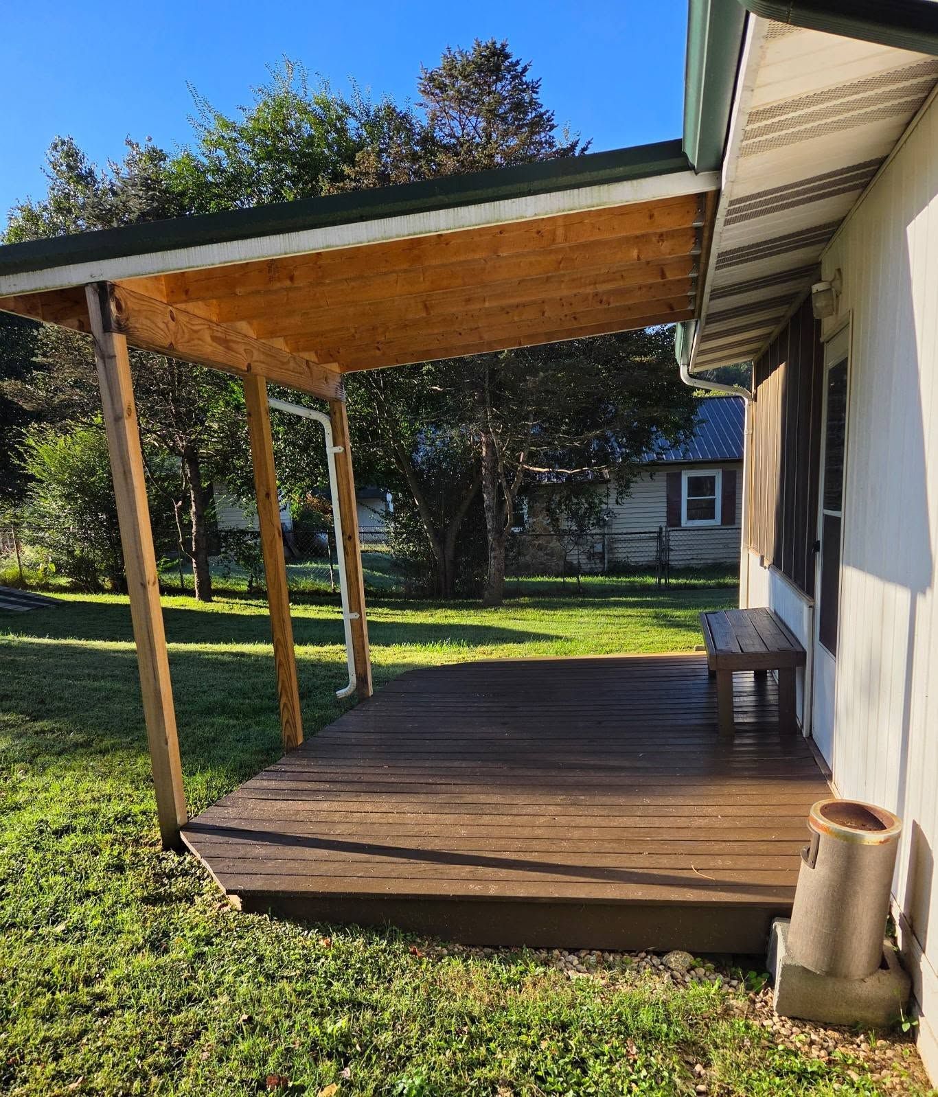 Wooden deck with roofed patio beside a white house, overlooking a grassy yard and trees.