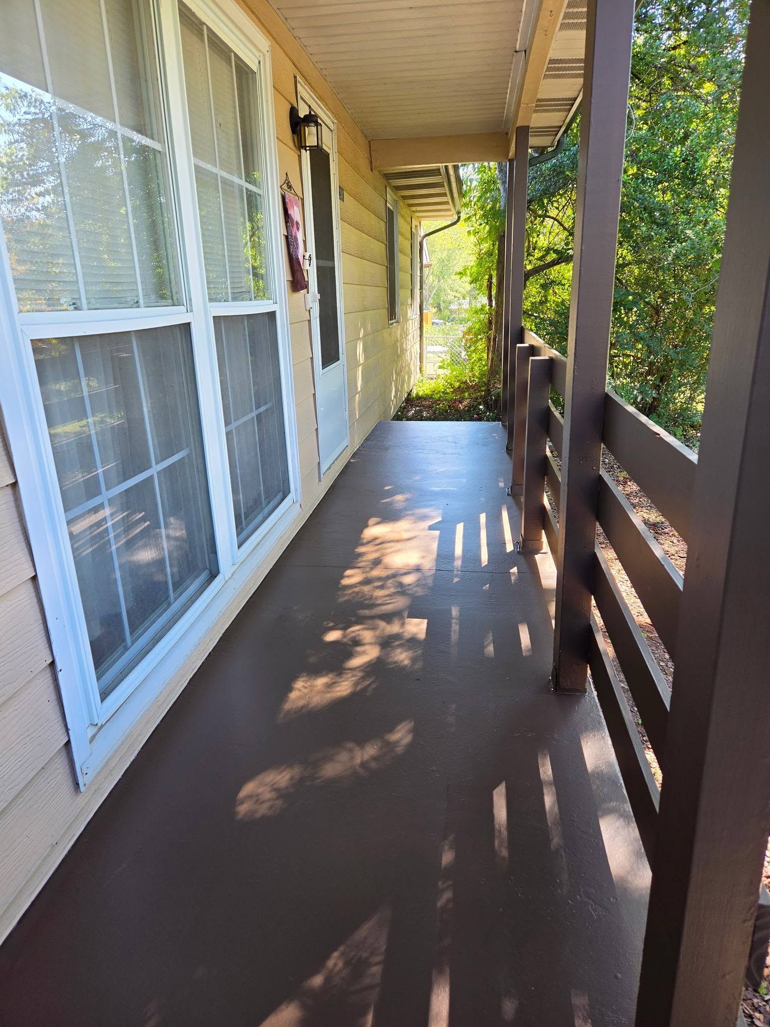 Sunlit apartment porch with brown floor, white siding, windows, and wooden railing overlooking greenery