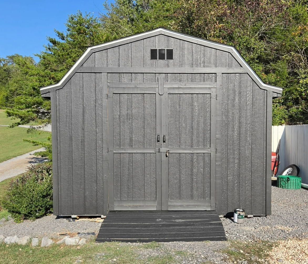 Gray garden shed with double doors and small windows, set on a gravel yard with trees behind it