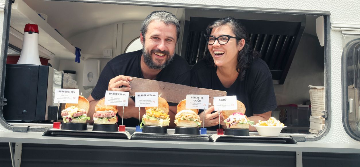 A man and a woman are standing in front of a food truck holding hamburgers.