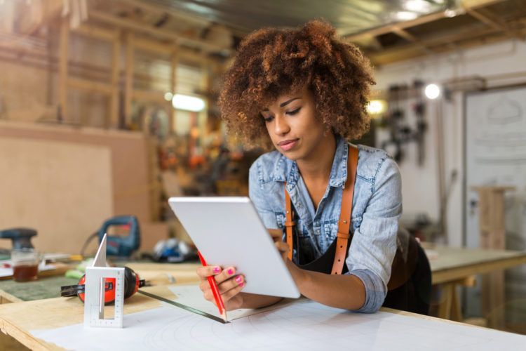 A woman is sitting at a table in a workshop using a tablet computer.