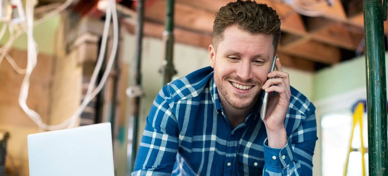 A man is sitting in front of a laptop and talking on a cell phone.