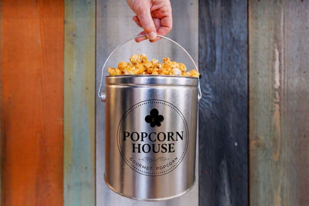 Hand holding a silver popcorn bucket against a rustic wooden wall, filled with popcorn.