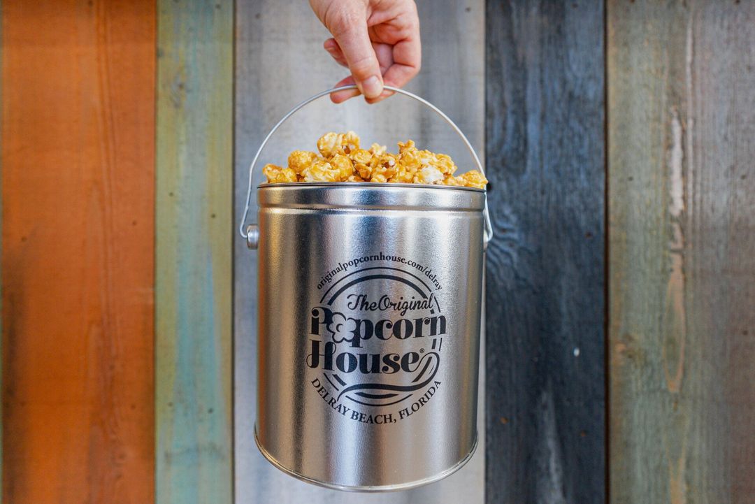 Hand holding a silver popcorn bucket against a rustic wooden wall, filled with popcorn.