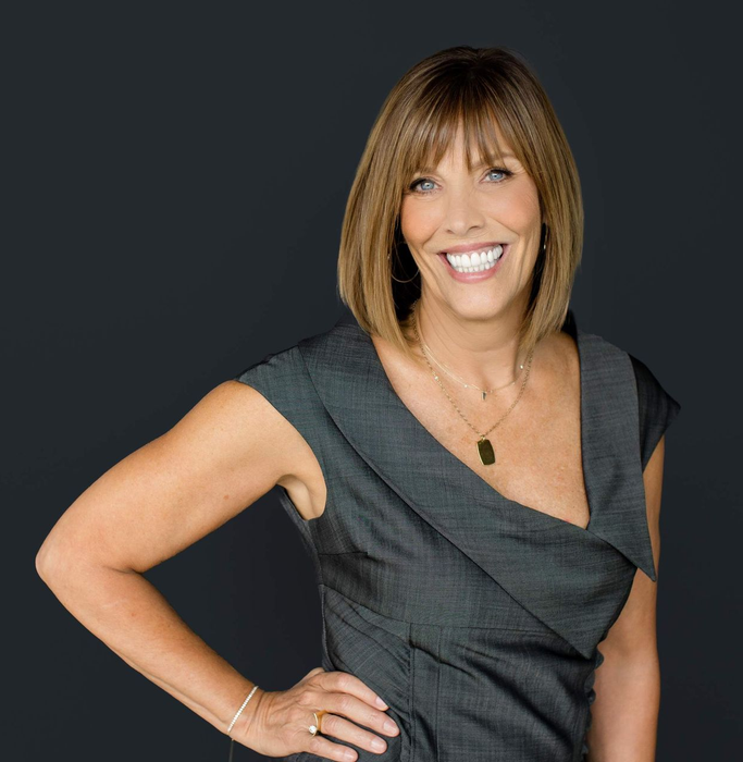 Woman with bob haircut smiles, wearing a gray dress, hand on hip, posing in front of a dark background.