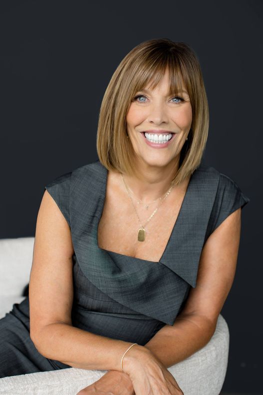 Woman with short brown hair, smiling, wearing a gray dress, sitting in a chair.
