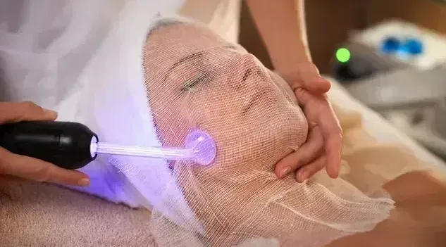 Person receiving facial treatment with a wand emitting a purple glow, face covered in mesh.