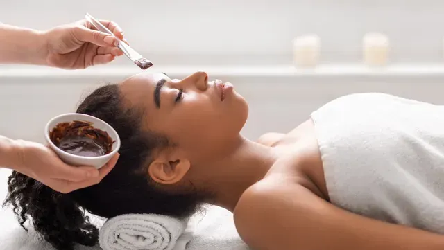 Person receiving a facial mask in a spa; a hand applies mask to forehead.