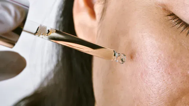 A pipette dispensing liquid onto a person's cheek near a blemish. Close-up, bright lighting.
