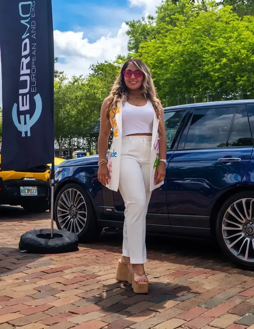 Woman in white outfit posing outdoors beside a dark SUV and Euro-made banner.