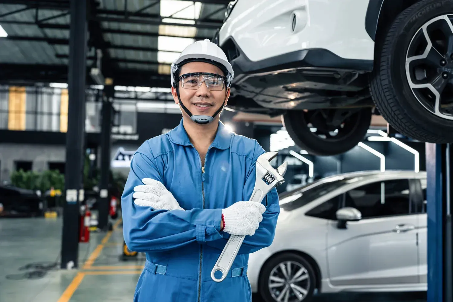 Mechanic in blue uniform holding a wrench under a lifted white car in an auto repair shop