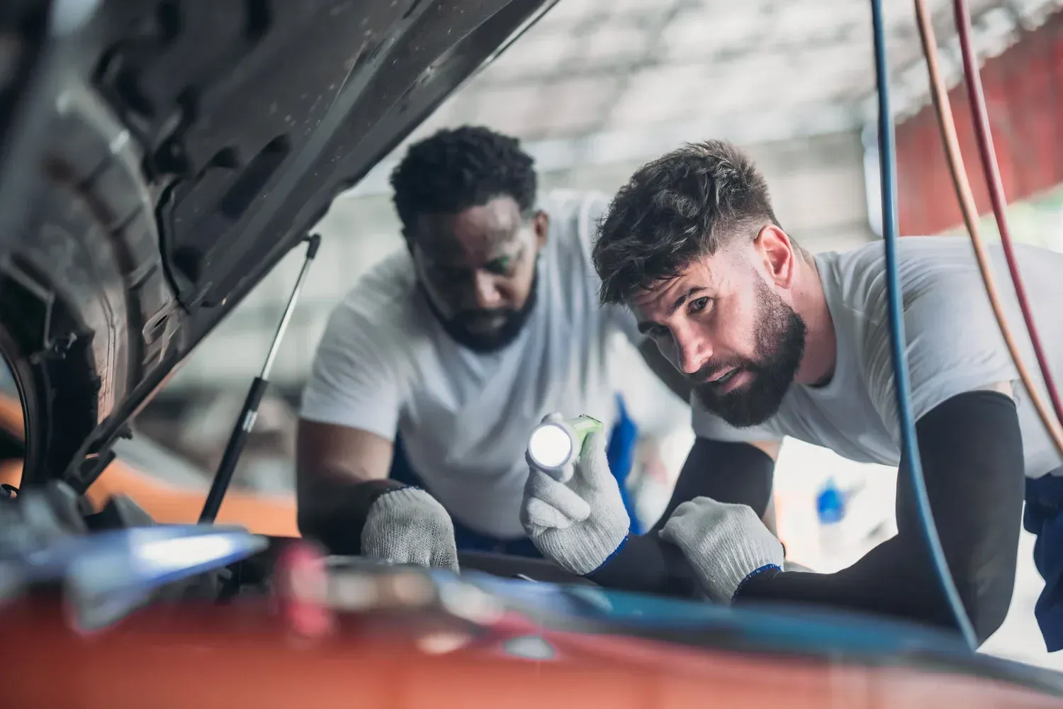 Two mechanics inspect a car engine in a garage, one holding a flashlight under the open hood.