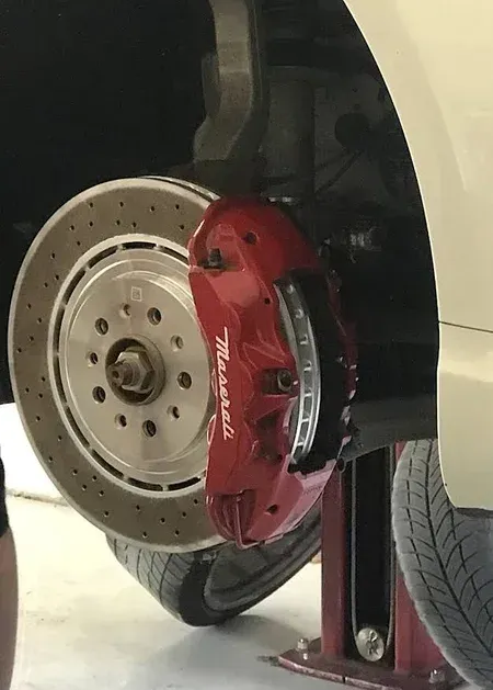 Mechanic removing a brake rotor, hands covered in grease, with a red car and oil drain bucket nearby