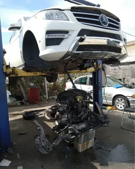White Mercedes SUV lifted above a wrecked black car in a repair yard, with debris below