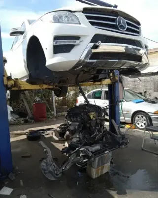 White Mercedes SUV lifted above a wrecked black car in a repair yard, with debris below