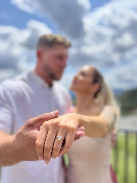 Engagement ring shown in foreground, with a couple softly blurred in the background outdoors under a cloudy sky