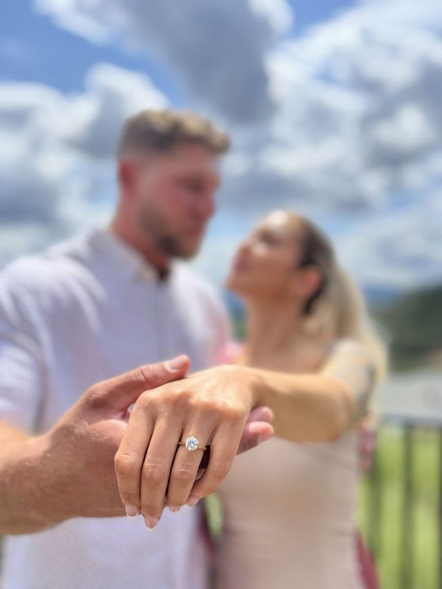 Engagement ring shown in foreground, with a couple softly blurred in the background outdoors under a cloudy sky