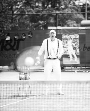 Een persoon in vintage tenniskleding met bretels houdt tennisballen vast op een tennisbaan, staand achter een ballenbak.