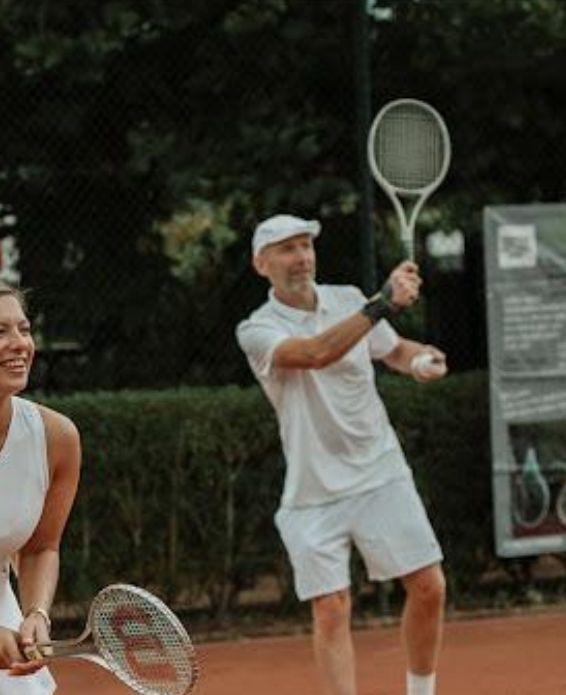  spelen tennis op een gravelbaan; een van hen staat op de voorgrond te glimlachen,