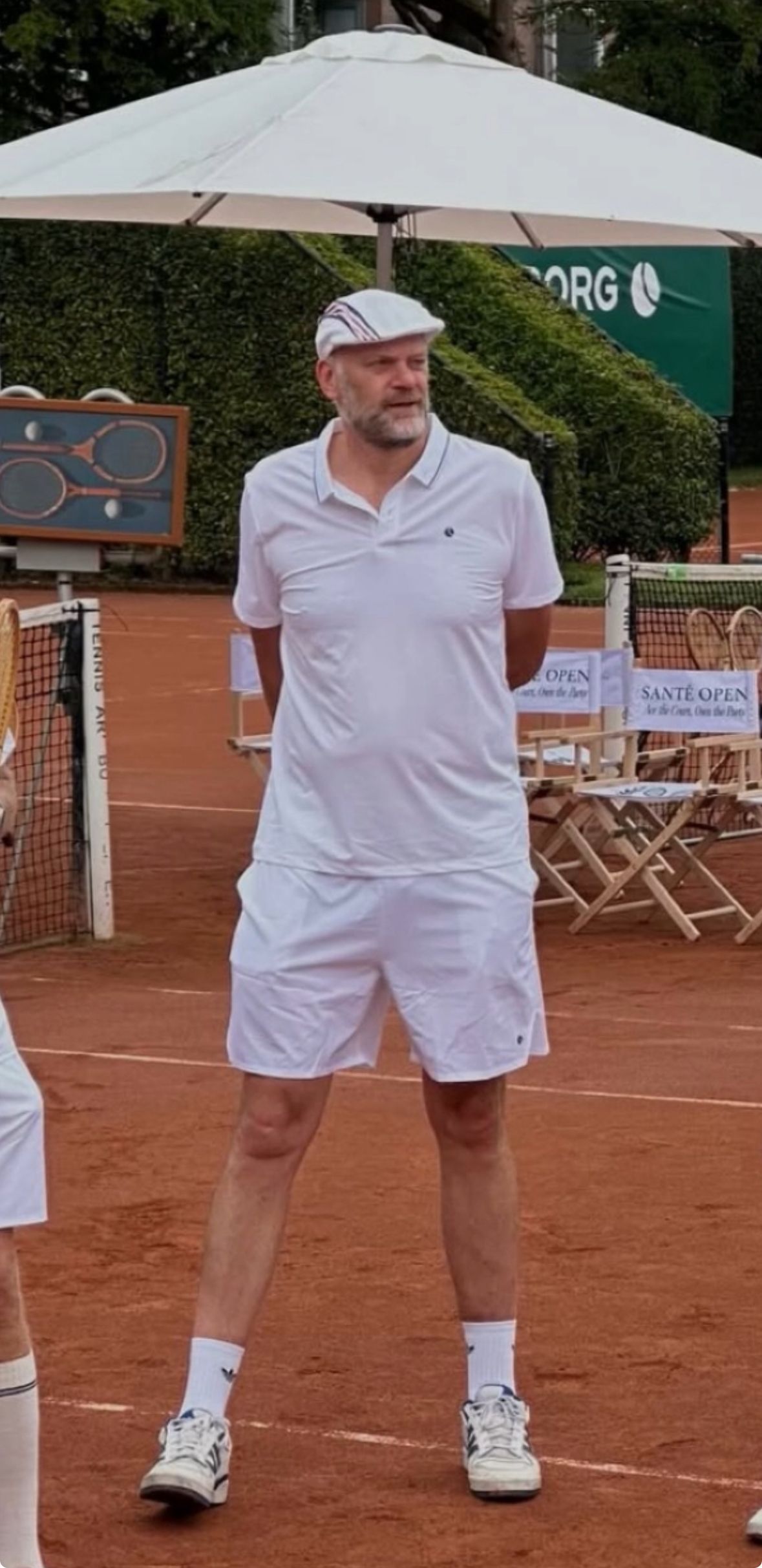 Een persoon in volledig witte tenniskleding en een platte pet staat op een rode graveltennisbaan