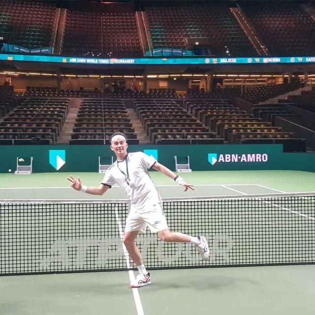 Een persoon in tenniskleding poseert speels op een tennisbaan in een stadion met ABN AMRO-reclame.