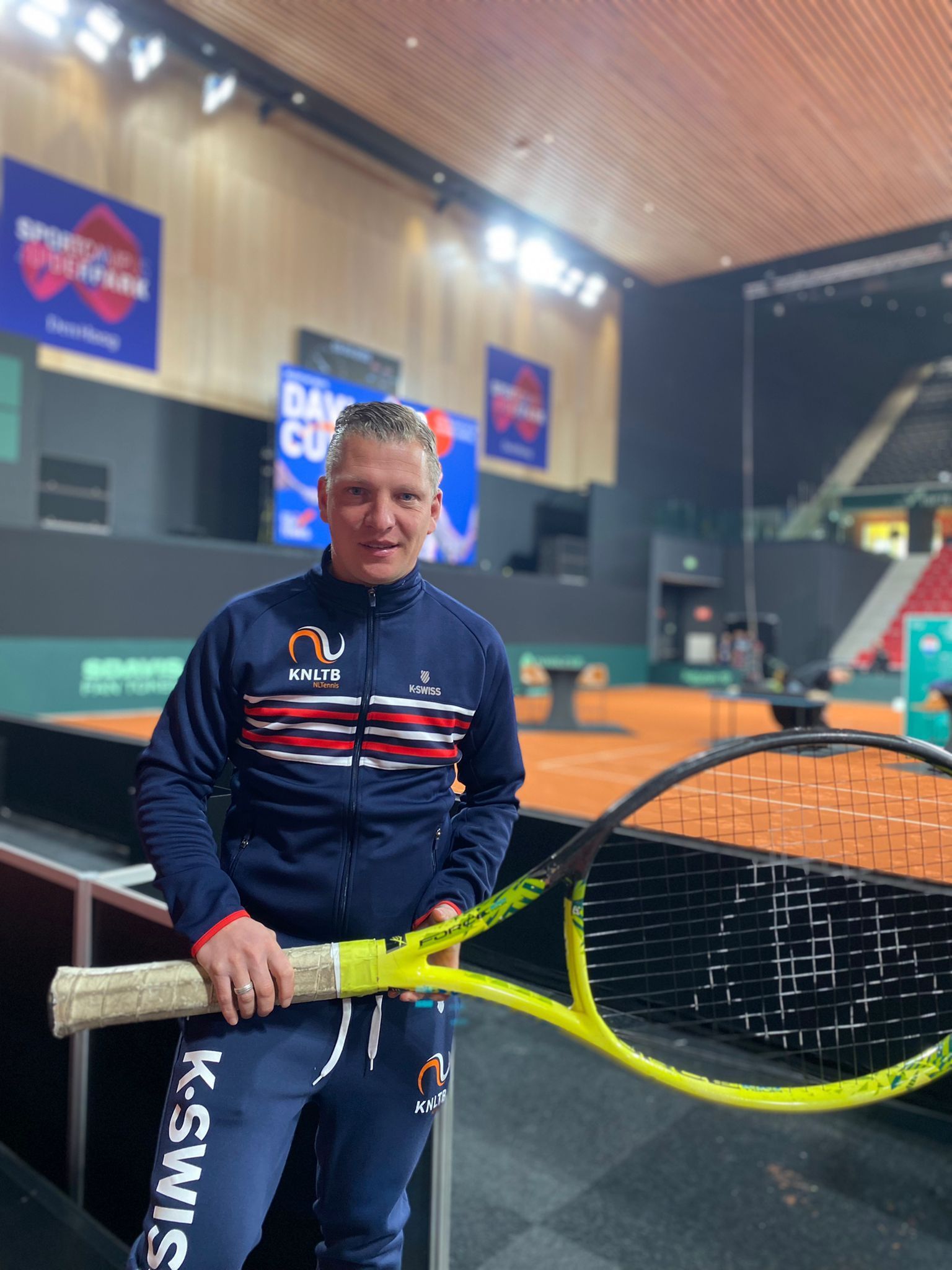 Een persoon staat in een tennisarena met een felgele tennisracket in zijn handen, voor een gravelbaan.