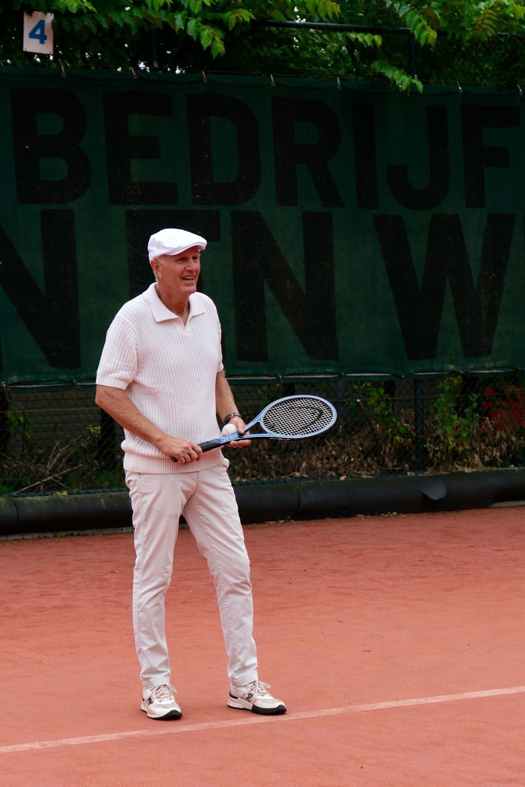 Een lachend persoon in een wit shirt, broek en pet, met een tennisracket in de hand, staat op een rode graveltennisbaan.