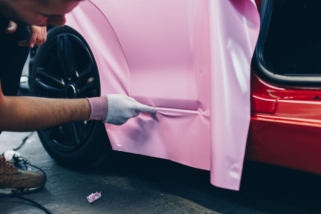 Person wrapping a car panel in pink vinyl beside a red vehicle wheel
