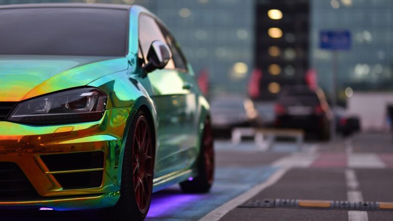 Shiny iridescent sports car parked on a city street at dusk, glowing with neon underlights.