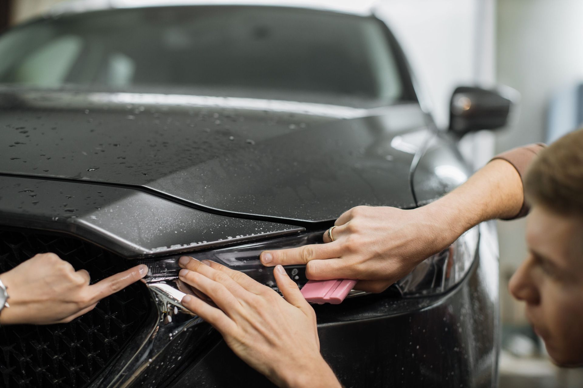 Hands cleaning a black car hood and headlight with water splashing in a car wash